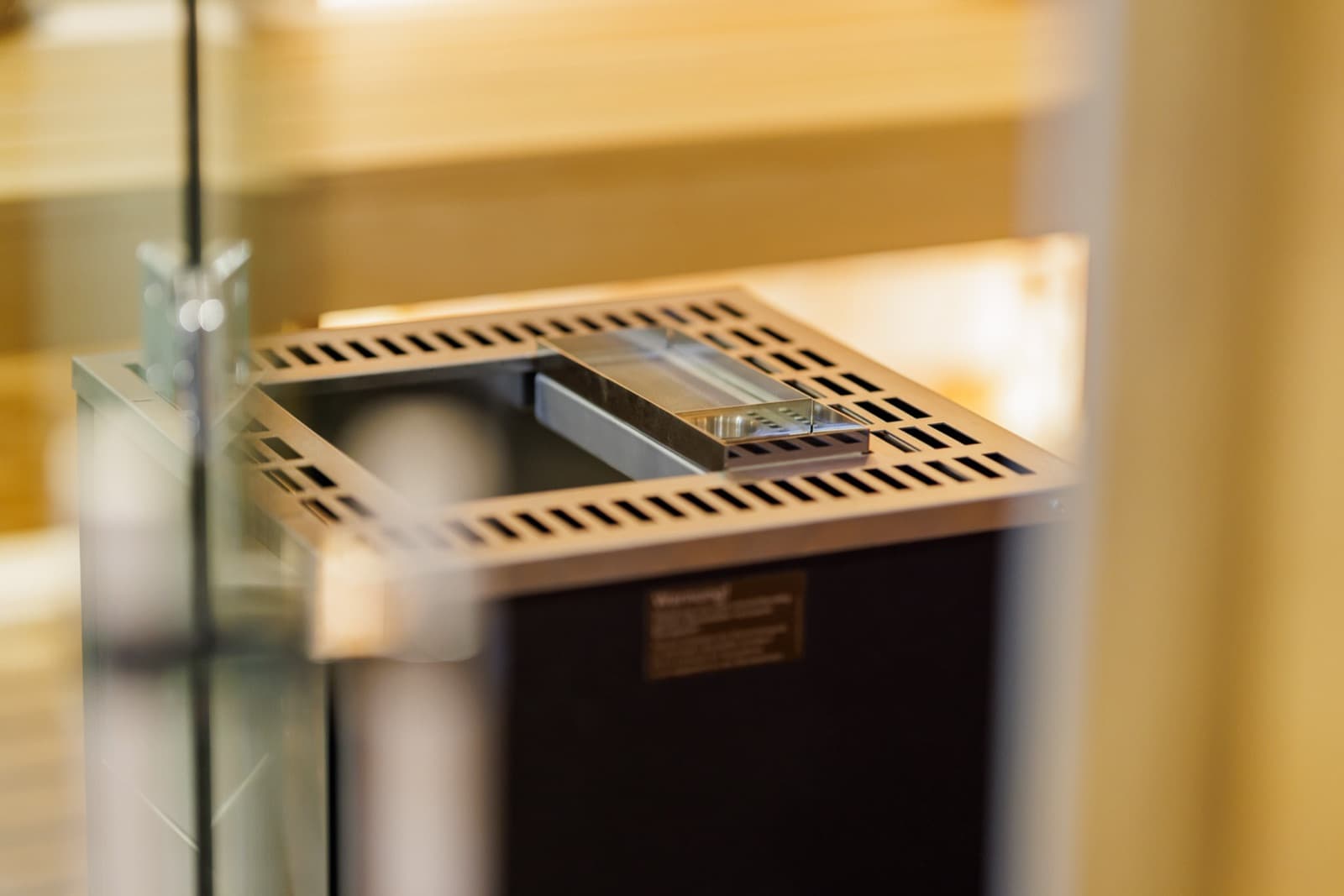 Nahaufnahme durch eine Glastür: Oberseite eines Elektro-Saunaofens mit geschlitztem Metallrahmen und flacher Metallschale, darunter ein Hinweisschild am dunklen Ofenkörper.