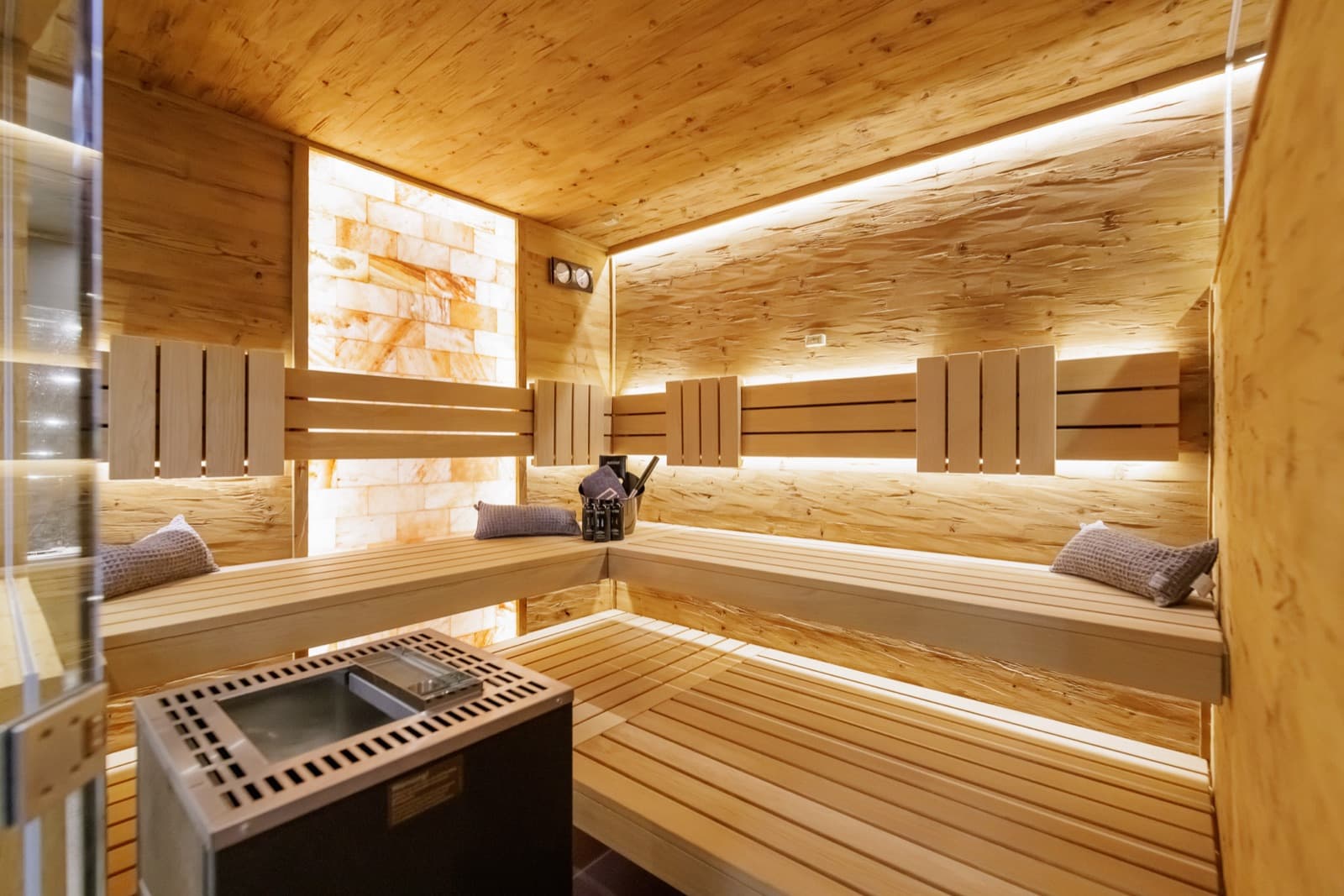 Saunaraum mit L-förmiger Bank aus hellem Holz auf zwei Ebenen, Rückenlehnen, hinterleuchteter Salzsteinwand, gehackter Holzwand mit indirektem Licht und einem Elektroofen neben dem Einstieg.