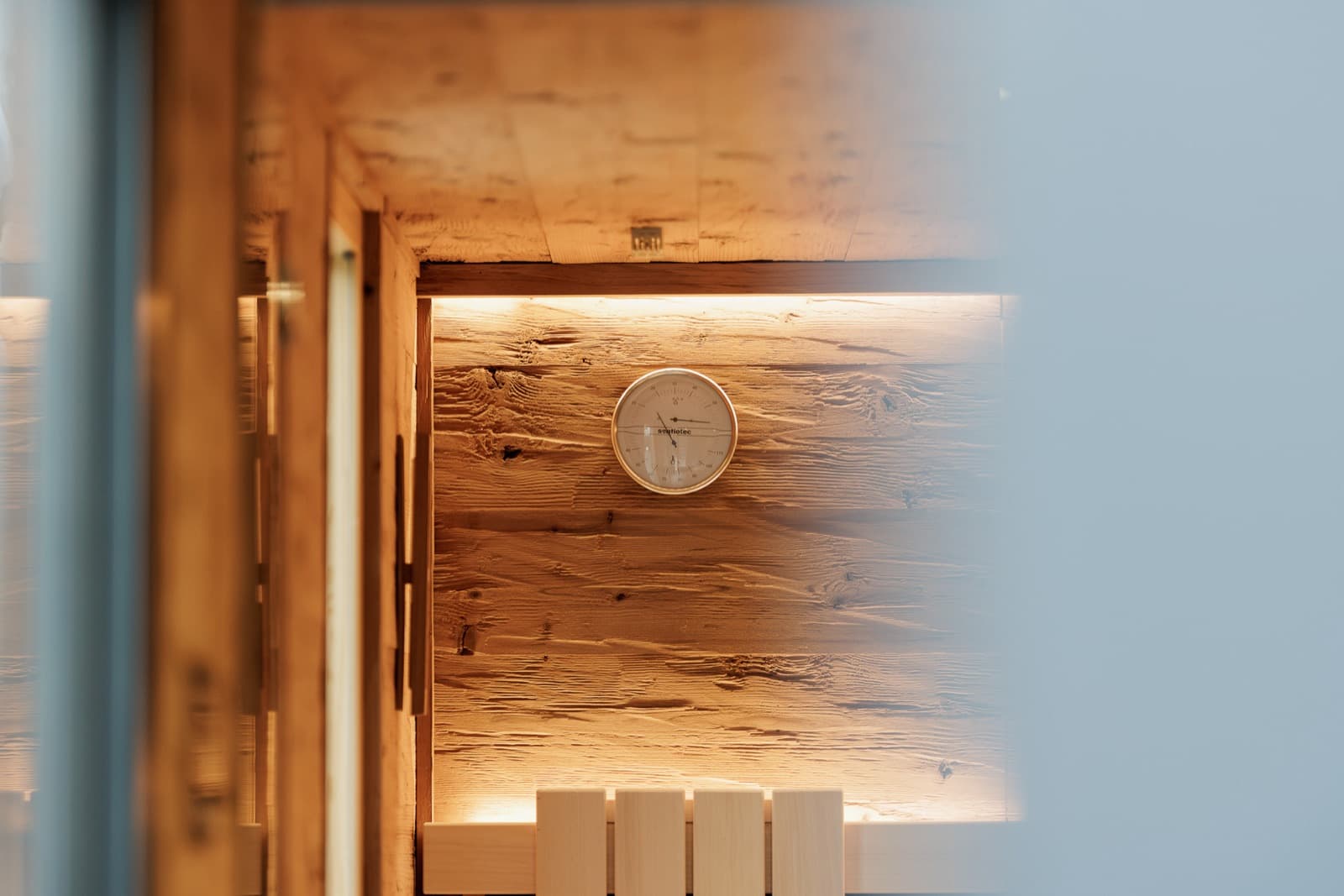 Blick durch eine geöffnete Glastür in einen Saunaraum: An der Holzwand in Altholzoptik hängt ein rundes Anzeigeinstrument, darüber verläuft eine indirekte Lichtlinie.