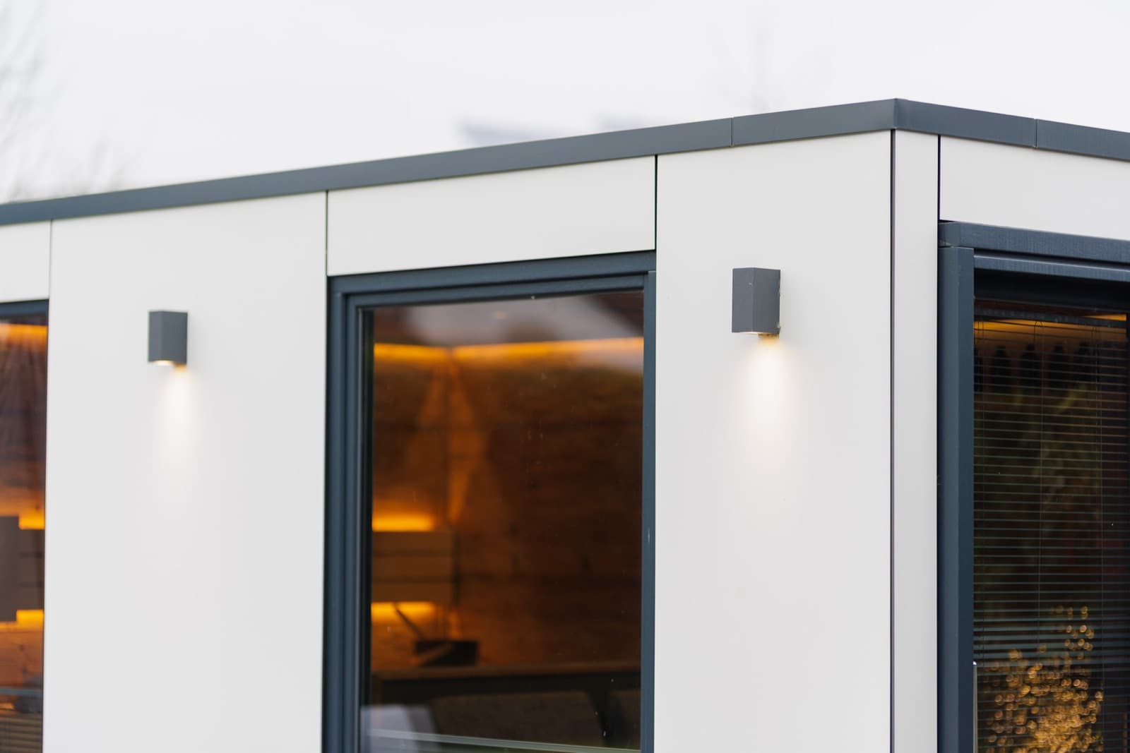 Ausschnitt einer Spa-Sauna mit heller Plattenfassade in der Dämmerung: Durch das Fenster in der Mitte ist der warm beleuchtete Saunaraum zu sehen, hinter der Scheibe rechts die Lamellen einer Jalousie.