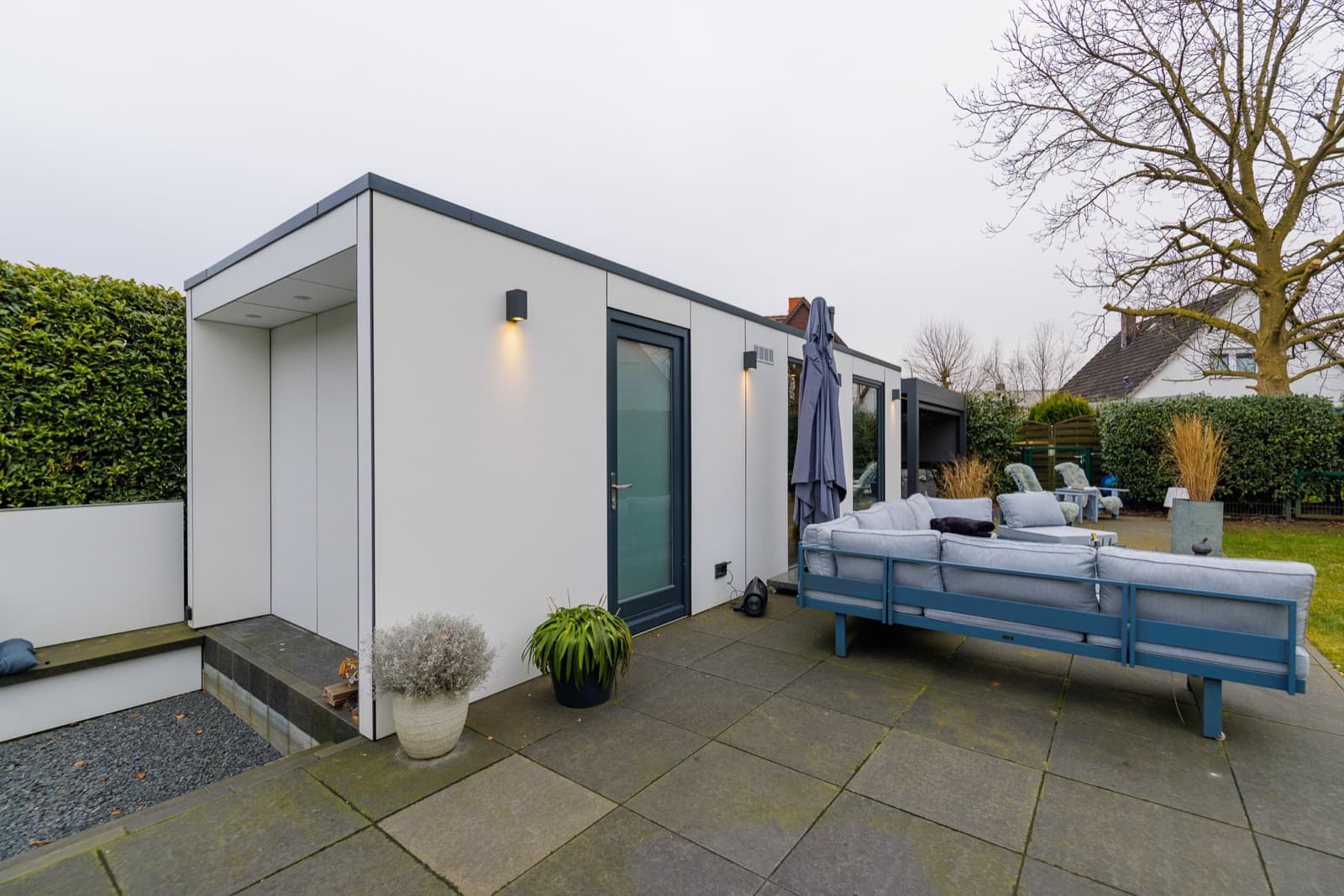 Weiße Spa-Sauna mit überdachtem Eingang und Glastür an einer Terrasse aus Steinplatten, rechts Loungemöbel, links eine hohe Hecke, im Hintergrund ein kahler Baum und Nachbarhäuser.