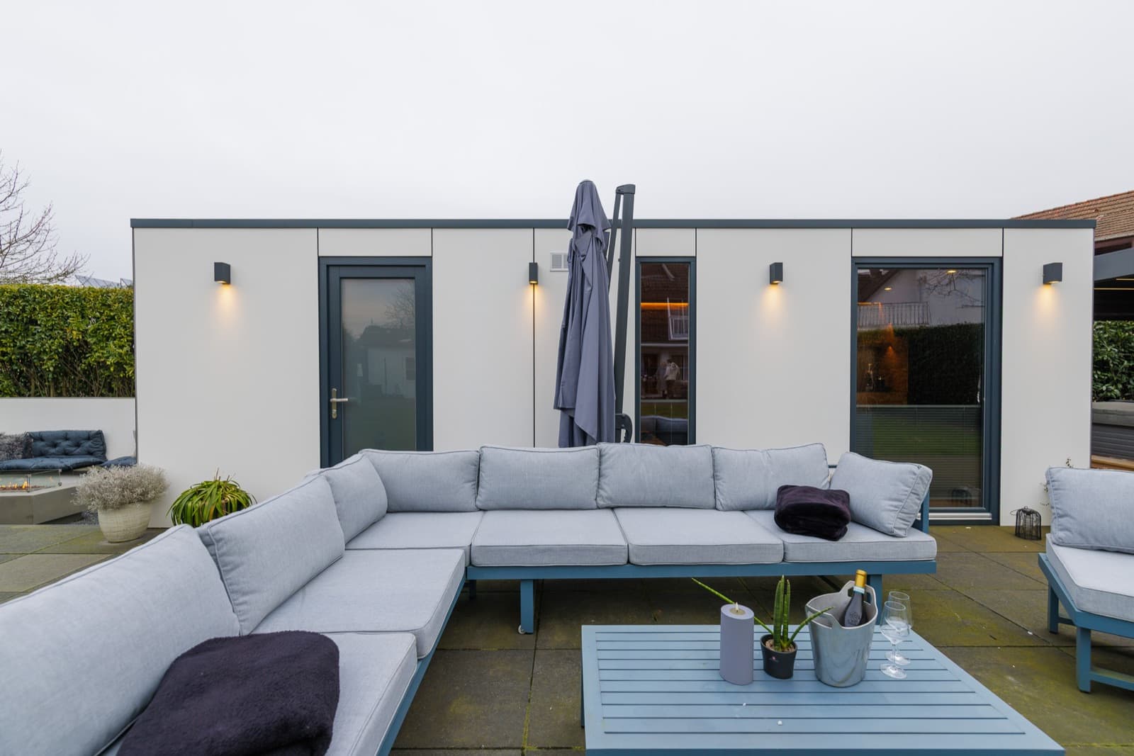 Helle Fassade einer Spa-Sauna mit vier kleinen Wandleuchten, die nach unten strahlen; davor eine Terrasse mit Loungemöbeln.