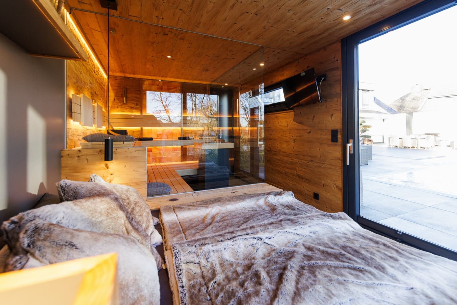 Ruheraum einer Spa-Sauna mit Liegefläche und Felldecken, Wandverkleidung in Altholzoptik, bodentiefer Glastür zur Terrasse und Saunaraum hinter einer Glaswand.
