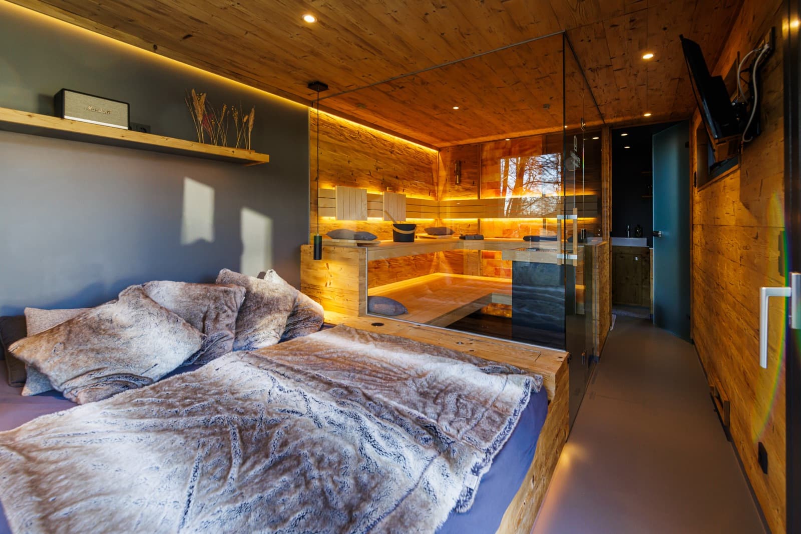 Ruheraum einer Spa-Sauna mit Bett und durchgehendem grauem Boden; hinter einer Glaswand der Saunaraum mit dunklem Boden und Holzbänken.