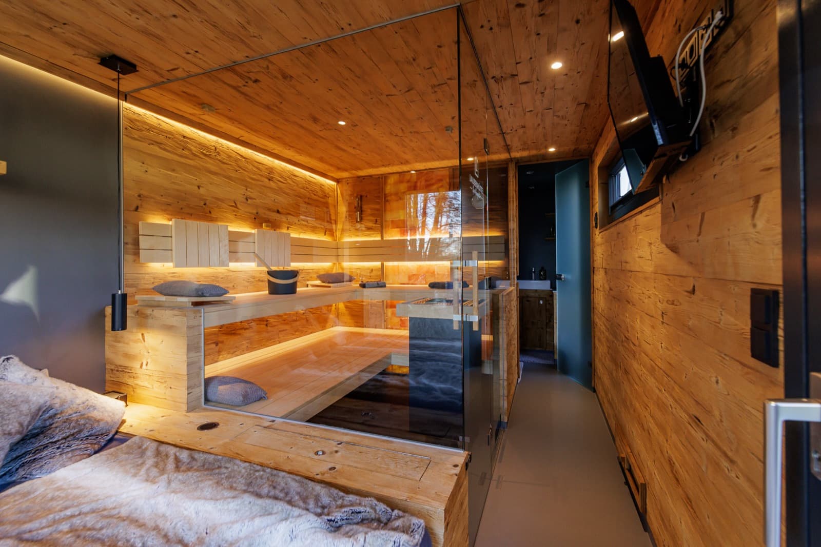 Ruheraum einer Spa-Sauna: vorn links eine Liegefläche mit Felldecken, in der Mitte der Saunaraum hinter einer Glaswand, rechts ein freier Durchgang entlang der Holzwand.
